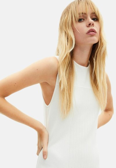 Vestito bianco senza maniche con texture a coste, collo alto e spacchi laterali. Capelli biondi lunghi e lisci che creano un contrasto con il tessuto.