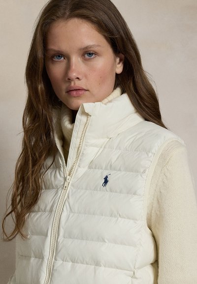 Jeune femme aux longs cheveux bruns portant un gilet matelassé blanc sur un pull crème, faisant face à l'avant avec une expression neutre.