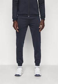 Tumši zili sporta bikses ar locītiem galiem un sānu kabatām, gludas tekstūras, kopā ar baltām sporta kurpēm.