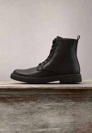 Bottine en cuir noir à bout rond, lacets à l’avant, languette au talon et semelle en caoutchouc plate reposant sur une surface en bois.