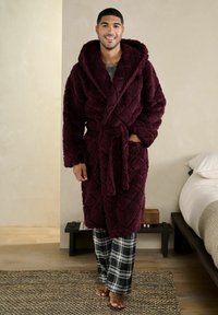 Mand står barfodet i soveværelset iført en vatteret burgunderrød hættebadekåbe og sort-hvide ternede pyjamasbukser, smilende.