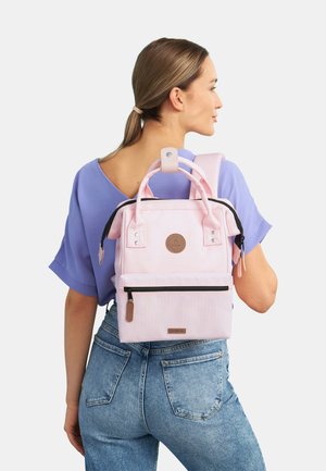 Petit sac à dos rose avec deux poignées supérieures et des bretelles réglables, comportant une poche zippée à l'avant et des accents en cuir marron.