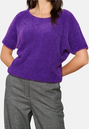 Kobieta w jasnofioletowym, puszystym swetrze z krótkim rękawem i szarych, kraciastych spodniach, ręce w kieszeniach, na jednolitym tle.