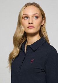 Marinblå pique-t-shirt i textil, med krage, tre knappar och en liten röd logotypbroderi på bröstet.