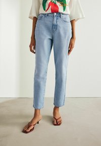 Jeans din denim de culoare albastru deschis, cu croi drept și ușoară decolorare, asortați cu sandale plate roșii. Are buzunare frontale și fermoar.