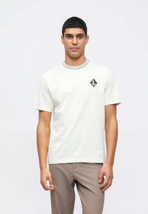 T-shirt à manches courtes de couleur crème en coton, avec un col rond bordé de gris et un logo en forme de diamant noir sur la poitrine gauche.