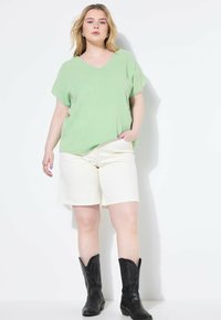 Top de cuello en V corto de manga corta en verde claro y texturizado, combinado con pantalones cortos crema que presentan un ajuste relajado y longitud estándar hasta el muslo. Botas de vaquero negras completan el look.