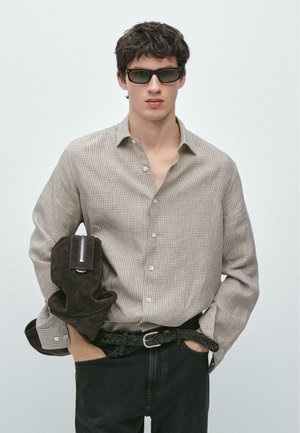 Jeune homme portant des lunettes de soleil, une chemise beige à carreaux, une ceinture noire tressée et tenant un sac en daim brun foncé sur un fond uni.