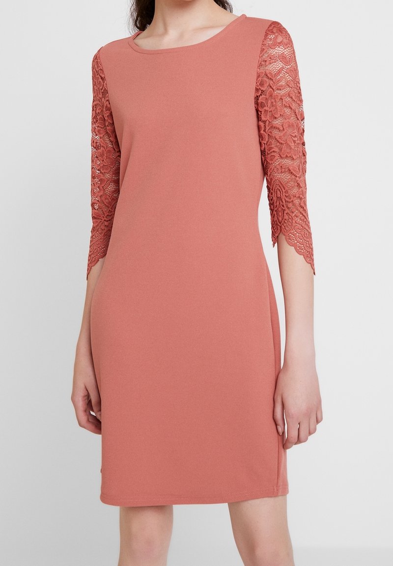 Femme portant une robe rose poudrée longueur genou avec manches trois-quarts en dentelle florale transparente et encolure ronde sur un fond uni.