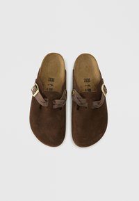 Καφέ σουέντ τσόκιο παπούτσια με φαρδιά λωρίδα και αγκράφα σε χρυσό τόνο, ανοιχτή πλάτη και ανάγλυφη σόλα που αγκαλιάζει το πόδι. Λείος υφές με απαλή φινίρισμα.