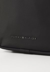 Crna najlonska torba s teksturiranim površinom i utonulim logom "TOMMY HILFIGER" u srebrnoj boji. Ima zaobljene rubove i izdržljivu konstrukciju.
