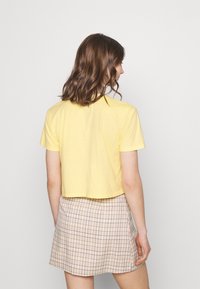 Žluté zkrácené tričko vyrobené z měkké tkaniny, s krátkými rukávy. V kombinaci s béžovou kostkovanou sukní s jemným mřížkovým vzorem.