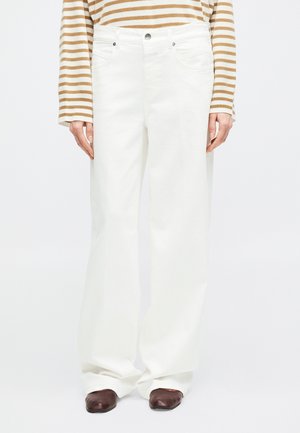 Personne portant un pantalon blanc taille haute à jambes larges, des chaussures marron à bout carré, et un haut à manches longues beige et blanc rayé.