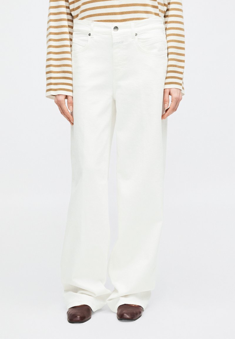 Personne portant un pantalon blanc taille haute à jambes larges, des chaussures marron à bout carré, et un haut à manches longues beige et blanc rayé.