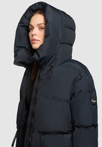 Navy blue puffer jacket with a large hood, featuring a black zip closure and logo patch on the sleeve. Smooth texture and lightweight design.