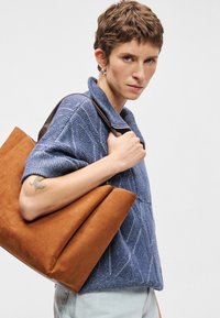 Καφέ σουέτ τσάντα tote με δομημένο σχέδιο, που διαθέτει τριγωνικό σχήμα και αντίθετη δερμάτινη λαβή. Μοντέλο που φοράει μπλε υφασμάτινο πουλόβερ.