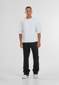 Witte t-shirt met korte mouwen, gecombineerd met zwarte jeans en witte schoenen. Het t-shirt heeft een losse pasvorm en de jeans zitten iets losjes.