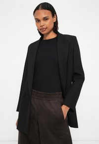 Sacou negru personalizat, cu rever ascuțit, purtat peste un top negru cu textură ribbed, asortat cu pantaloni negri din material elastic cu talie elastică. Textură de țesătură netedă.