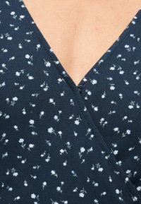 Ύφασμα σε ναυτικό μπλε με μικρό λευκό φλοράλ μοτίβο και ντεκολτέ σε σχήμα V που επικαλύπτεται στο κέντρο πάνω σε ανοιχτόχρωμο δέρμα.