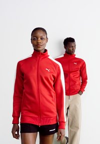 Rød zip-up jakke med hvide detaljer, lavet af blødt materiale. Modellen foran har den på over sorte shorts. Bagmodellen har en matchende jakke på.