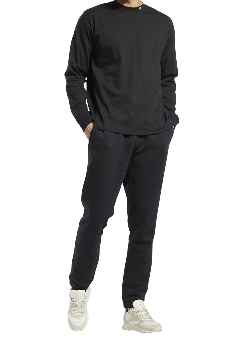 Schwarzes Langarmshirt mit Rundhalsausschnitt, kombiniert mit schwarzen Jogginghosen. Dazu werden hellfarbene Sneakers mit einer strukturierten Sohle getragen.