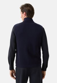 Granatowa wełniana kamizelka z wysokim kołnierzem i bezrękawnikowym designem. Widok z tyłu pokazuje gładką teksturę i proste, czyste linie.