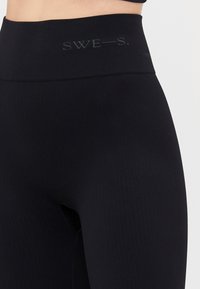 Nærbilde av svarte ribbestrikkede høytalende leggings med "SWE—S." logoen på midjebåndet.