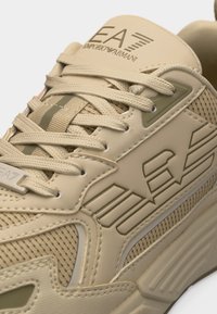 Μπεζ sneaker Emporio Armani με υφή από διχτυωτό ύφασμα, χοντρή σόλα, λογότυπο "EA7" δεξιά στην πλευρά και στη γλώσσα, και μπεζ κορδόνια περασμένα από μεταλλικές τρύπες.