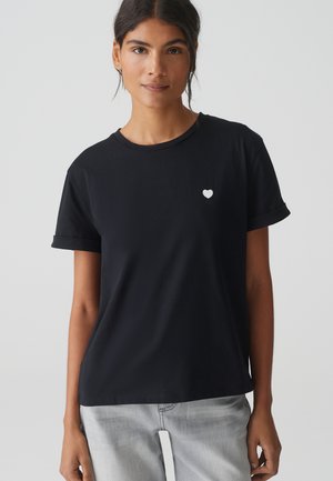 Donna con capelli scuri che indossa una maglietta nera con un piccolo emblema a cuore bianco sul lato sinistro del petto e jeans grigio chiaro, sfondo neutro.