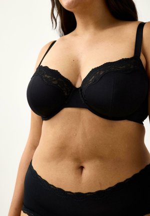 Donna che indossa un reggiseno nero con pizzi e mutandine coordinate, corti al collo e alle cosce, su uno sfondo chiaro e semplice.