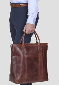 Braune Leder-Tote-Tasche mit robusten Griffen, versehen mit einer Fronttasche und sichtbaren Steppnähten, getragen von einer Person in formeller Kleidung.