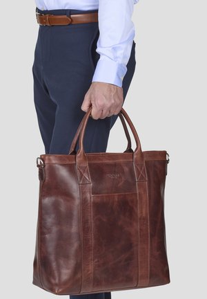 Brown leather tote bag with sturdy handles, featuring a front pocket and visible stitching details, carried by a person in formal attire.