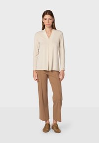 Beige Langarmoberteil mit V-Ausschnitt und subtilen Faltendetails, kombiniert mit hellbraunen weit geschnittenen Hosen und braunen Wildledersneakern.