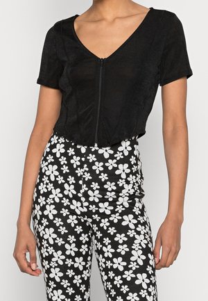 Kobieta w czarnej bluzce na krótki rękaw z suwakiem, do której dobrano czarne spodnie z białym kwiatowym wzorem.