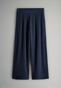 Pantalons évasés côtelés bleu marine avec une taille élastique, suspendus devant un fond gris clair uni.