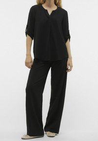 Blusa nera a maniche lunghe con scollo a V, in tessuto texturizzato, abbinata a pantaloni neri larghi e svasati. Mules beige con dettaglio a fiocco.
