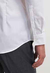 White dress shirt with a smooth texture, buttoned cuffs, and a curved hem. Worn with dark textured trousers.