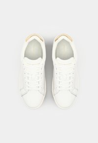 Baskets blanches avec une finition en cuir lisse, présentant un accent beige clair au niveau du talon. Design à lacets avec logo en relief sur la languette.