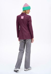 Fioletowa, teksturowana marynarka z klapkami, z metką z tyłu, zestawiona z jasnoszarymi, szerokimi jeansami i czapką z daszkiem w zielono-różowe wzory.