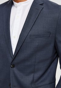 Mørkeblå dressjakke med teksturert stoff, enkelt knappelukking og brystlomme. Har en hvit skjorte under.