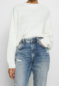 Personne portant un pull blanc en grosse maille rentré dans un jean bleu clair délavé et déchiré taille haute, avec la main dans la poche.