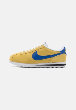 Baskets basses jaunes avec le swoosh Nike bleu, lacets blancs et semelle blanche avec une bande noire, vues de côté gauche sur fond blanc.