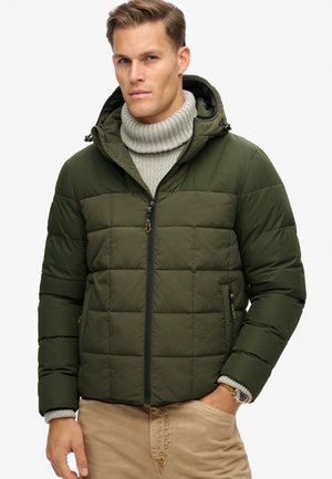 Chaqueta puffer de color verde oliva con cierre frontal de cremallera, capucha y diseño acolchado. Presenta bolsillos laterales y un suéter gris de canalé debajo.