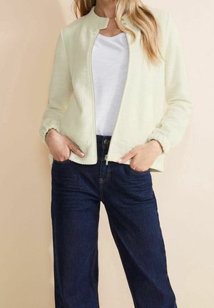 Woman wearing a cream textured zip-up jacket, white T-shirt, and dark blue jeans with hands in pockets against a beige background.
