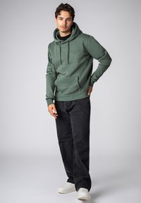 Groene hoodie met trekkoorden, kangoeroezak en gedrukte tekst op de mouw, gecombineerd met zwarte jeans en witte sneakers.