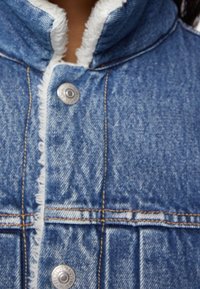 Jeansjacka med fransad kragsömn, silverfärgade tryckknappar och synlig sömnadsdetalj. Predominant blå med strukturerad tyg.