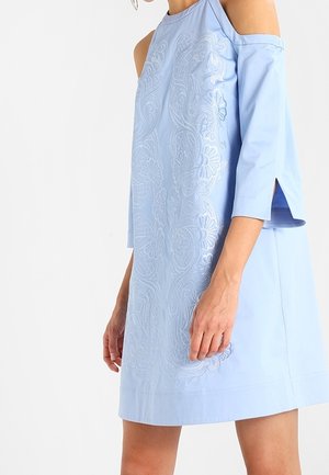 Mujer con vestido azul claro de hombros descubiertos, mangas tres cuartos y bordado floral en el frente, posando frente a un fondo blanco.