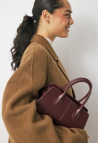 Burgundijas ādas rokassoma ar divām izliekām rokturiem, strukturētu formu un redzamām šuvēm, turēta pretim brūnai manti, kurai ir apjomīgas piedurknes.