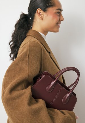 Bordeaux leren handtas met twee gebogen handvatten, gestructureerde vorm en zichtbare stiksels, vastgehouden tegen een bruine jas met volumineuze mouwen.