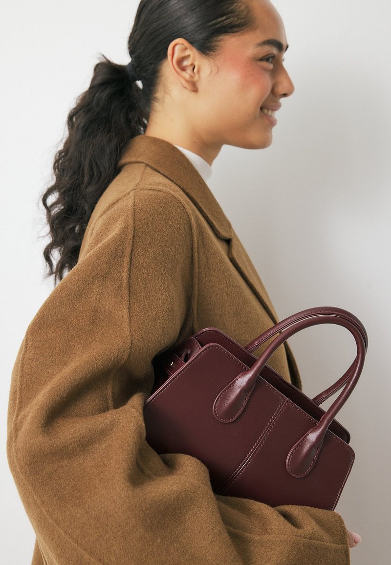 Burgundijas ādas rokassoma ar divām izliekām rokturiem, strukturētu formu un redzamām šuvēm, turēta pretim brūnai manti, kurai ir apjomīgas piedurknes.